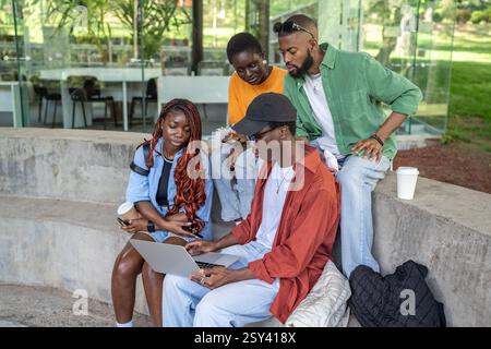 Black friends sitting on park bench with laptop, staring at screen. Working on coursework project Stock Photo