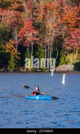 Solo female kayaker in a blue boat on Lac Stukely amid vivid autumn color, Parc national du Mont-Orford, Québec, Canada. Stock Photo