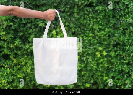Blank white tote bag canvas fabric with handle mock up design. Close up of woman hand holding eco or reusable shopping bag on green leaves background. Stock Photo