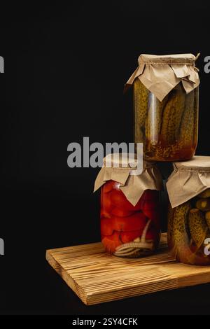 Three jars of homemade canned pickles, tomatoes, and sweet peppers are stacked on a wooden board. The jars are sealed with paper tops, showcasing the Stock Photo