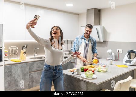 Happy young couple taking selfie with smartphone while preparing healthy food in modern kitchen Stock Photo