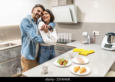 Young couple hugging in their modern kitchen, enjoying a romantic breakfast of strawberries, croissants, and donuts Stock Photo