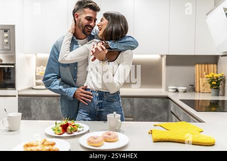 Young couple hugging and laughing in their modern kitchen, enjoying a romantic moment together with breakfast ready on the counter Stock Photo