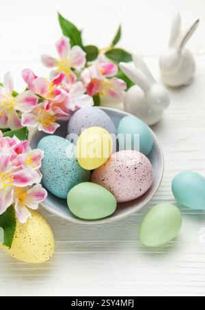 Colorful pastel easter eggs in a bowl with spring flowers and porcelain bunnies celebrating easter holiday on white wooden background Stock Photo
