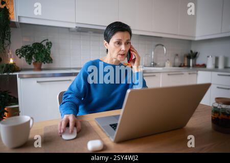 Focused businesswoman working remotely, managing tasks on laptop and smartphone in home office Stock Photo