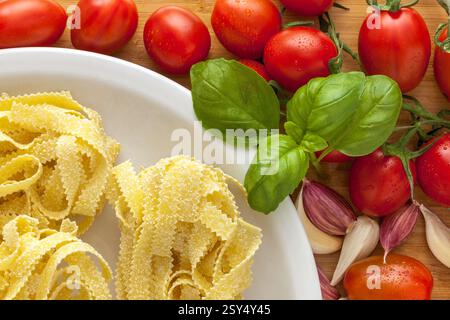 plate of durum wheat pasta, egg tagliatelle format, on wooden cutting board with ripe tomatoes, fresh basil leaves and garlic cloves Stock Photo