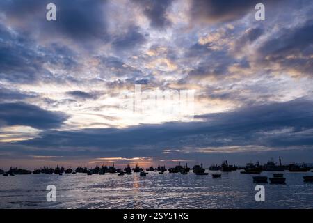 Many fishing boats are anchored off the beach of Mui Ne, Mũi Né, the sun is setting behind dark storm clouds. Stock Photo