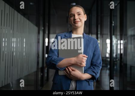 Confident young student, intern, or aspiring professional standing in a modern office hallway, holding a laptop. Concept of career growth, education Stock Photo