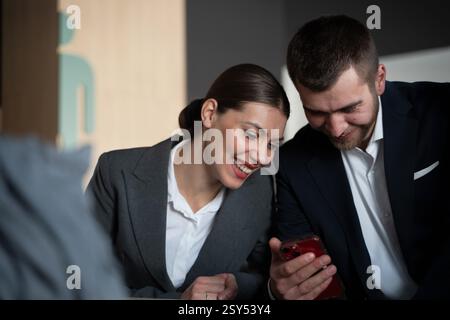 Two business professionals in formal attire laughing while looking at a smartphone screen in an office environment. Concept of teamwork, workplace Stock Photo