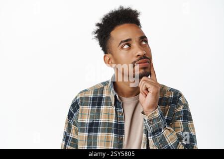 Close up of thoughtful african american man making decision, student thinking about smth, touch jaw and looking up, ponder choices, standing over whit Stock Photo