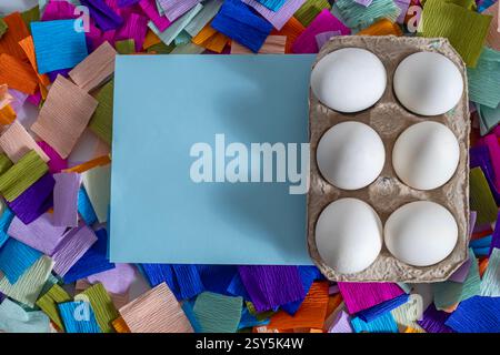 White eggs on a cardboard box with blank blue card and vibrant confetti pieces, perfect for holiday celebrations, Easter-themed messages and designs. Stock Photo