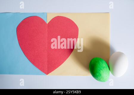 A colorful arrangement of a red paper heart with green and white eggs on a light blue and yellow background. Stock Photo