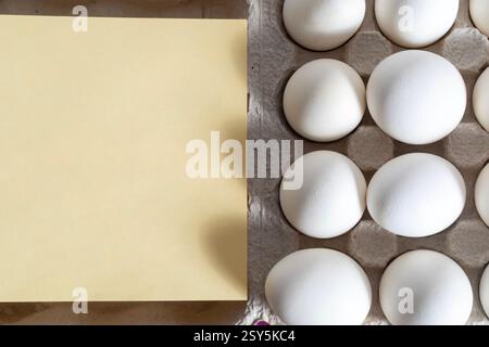 A dozen white eggs are stacked in a carton with blank yellow paper for design or text. Stock Photo