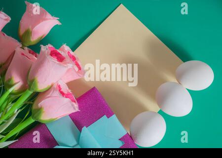 A flat lay composition with pink roses, wrapped gift, egg and blank card on colorful green background. Stock Photo