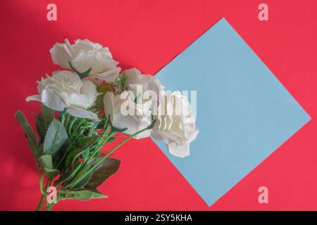 Top view of beautifully arranged artificial white roses with a blank card on a vibrant red background. Stock Photo