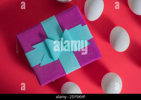 A vibrant purple gift box elegantly tied with a blue ribbon surrounded by white eggs, against a vibrant red background. Stock Photo