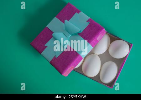A shiny purple gift box tied with a blue ribbon on a green background with white eggs. Perfect for surprise or unique gift ideas. Stock Photo
