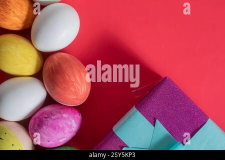 Easter eggs decorated in vibrant colors next to a wrapped gift box on a bold red background. holiday celebration. Stock Photo