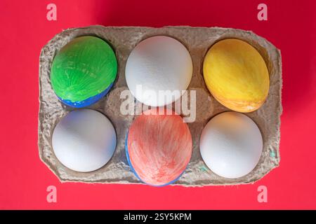 Top-down view of painted and empty chicken eggs in a carton with a vibrant red background. Stock Photo