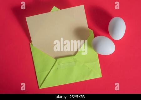 A bright and creative arrangement of a green envelope with paper and two white eggs placed on a vivid red background. Stock Photo