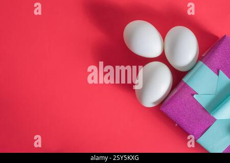 Three white eggs next to a vibrant, shiny, gift box with copy space for text, on a red surface. Stock Photo