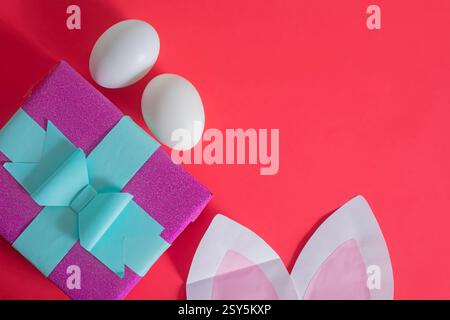 A vibrant Easter composition featuring a red background decorated with bunny ears, a colorful gift box and white eggs. celebration and seasonal joy. Stock Photo