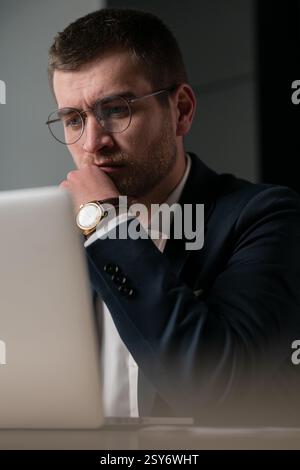 Vertical: Serious young businessman wearing glasses and a suit, deeply focused on his laptop. Concept of concentration, decision-making, remote work Stock Photo