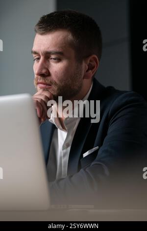 Vertical: Young entrepreneur or manager in a suit working on a laptop, deep in thought with a serious expression. Concept of corporate decision-making Stock Photo