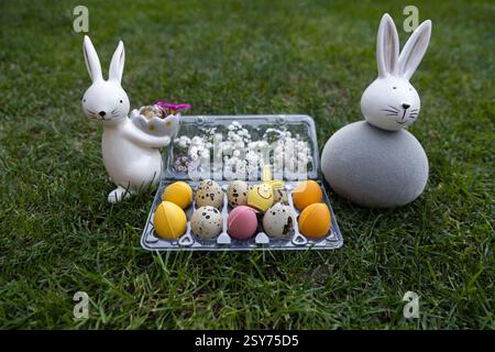 Two souvenir porcelain Easter bunny on a green lawn next to quail and multi-colored chocolate eggs. Home decor, festive Easter Stock Photo