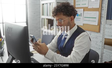 Young man focuses intently on smartphone screen while working at desk in modern office, surrounded by computer and bookshelves. Stock Photo
