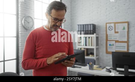 Middle-aged hispanic man in red sweater using tablet in modern office with computer and bookshelves enhancing digital productivity indoors. Stock Photo
