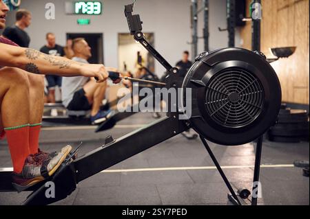 Close up side view. Sportive man doing exercises on rowing machine in the gym. Stock Photo