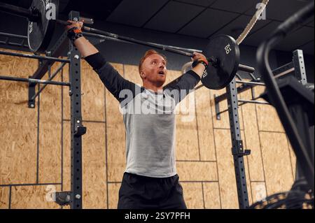 Standing with hands straight up. Strong man is lifting barbell in the gym. Stock Photo