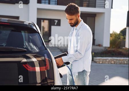 View from the side. Charging the vehicle. Young stylish man is with electric car at daytime. Stock Photo