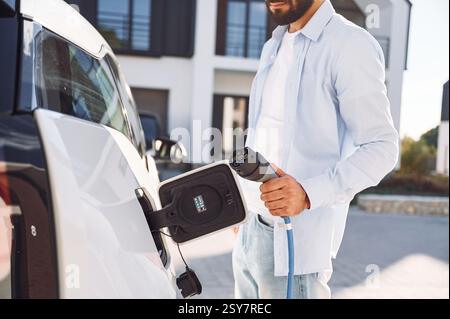 View from the side. Charging the vehicle. Young stylish man is with electric car at daytime. Stock Photo