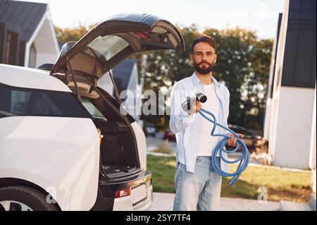 Holding yellow charging cable. Young stylish man is with electric car at daytime. Stock Photo