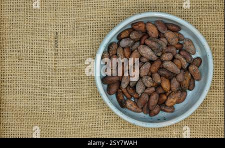 The bowl of cocoa beans. Cocoa beans in a handmade pale blue ceramic bowl on the jute background. The price and cost of cocoa beans. Top view. Stock Photo