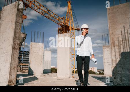 Talking by phone and holding document in hands. Man in uniform is working on the construction site. Stock Photo