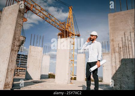 Talking by phone and holding document in hands. Man in uniform is working on the construction site. Stock Photo