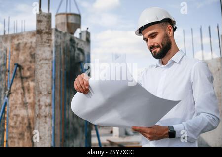 Standing and holding plan in hands. Man in uniform is working on the construction site. Stock Photo