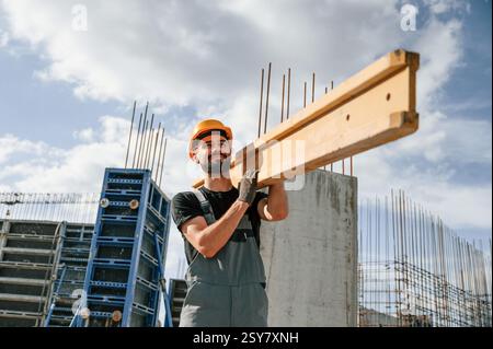 Carrying wooden plank. Man in uniform is working on the construction site. Stock Photo