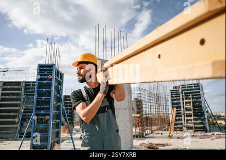 Carrying wooden plank. Man in uniform is working on the construction site. Stock Photo