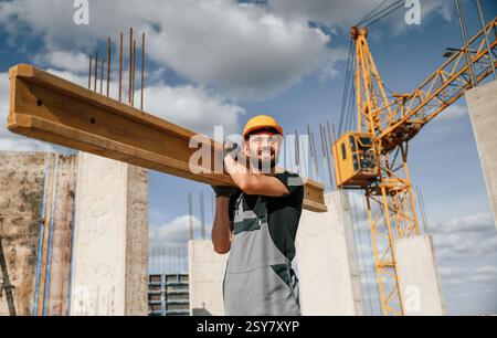 Holding and walking with wooden plank. Man in uniform is working on the construction site. Stock Photo