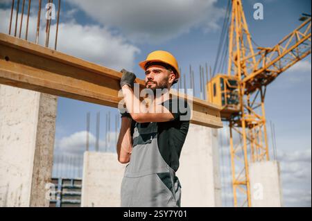 Holding and walking with wooden plank. Man in uniform is working on the construction site. Stock Photo