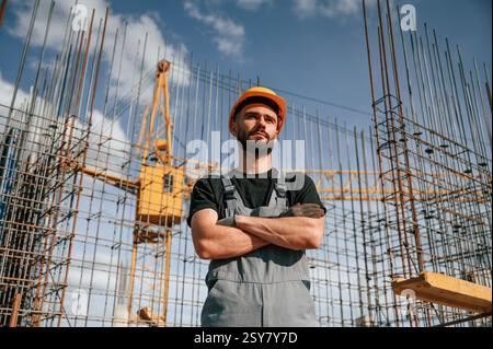 Standing against the crane with arms crossed. Man in uniform is working on the construction site. Stock Photo