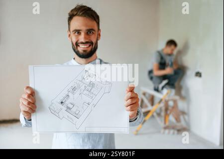 Holding plan of a room and smiling. Two men is making repairs in the apartment together. Stock Photo