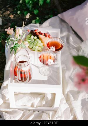 A charming setup featuring fruit, drinks, and flowers arranged on a white table outdoors during a summer picnic. Stock Photo