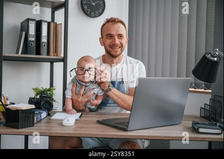 Big glasses. Sitting by the table with laptop. Father with his newborn son is indoors at home. Stock Photo