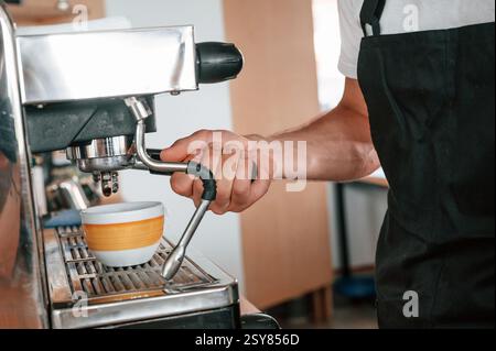 Using coffee machine. Cafe worker in white shirt and black apron is indoors. Stock Photo