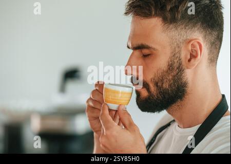 Side view. Tasting delicious coffee. Cafe worker in white shirt and black apron is indoors. Stock Photo